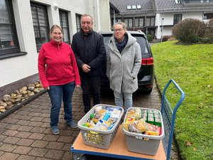 Dieter Gebauer und Elke Preuss übergeben zwei vor ihnen stehende Körbe mit Lebensmitteln an die Zuständige des Kreuztaler Mittagstisches, vor einem Haus und Auto stehend.